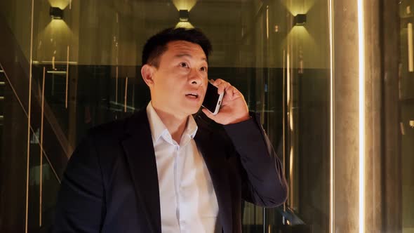 Asian Businessman Talking on Mobile Phone Rides in a Panoramic Elevator