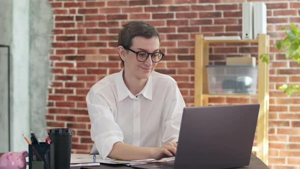 Smiling Businesswoman in Glasses and a White Shirt Works Behind a Laptop in the Office and Shows