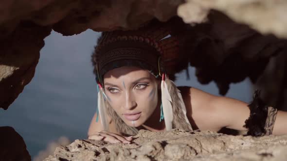 Strong Free Wild Woman with Tribal Ornamental Makeup and Feather Hat Hiding in Rocks and Hunting