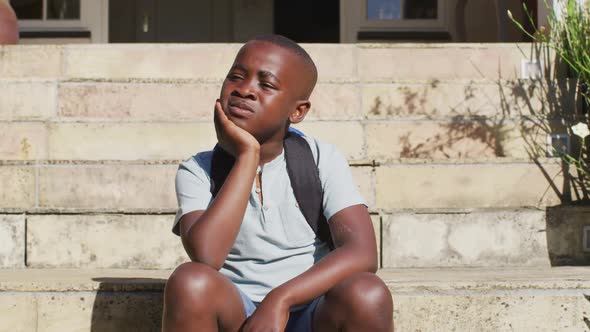 Sad african american boy wearing face mask sitting on stairs outdoors on a sunny day