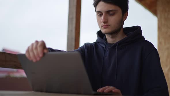 A Young Man is Typing on a Laptop While Sitting on a Terrace Against the Backdrop of a Picturesque