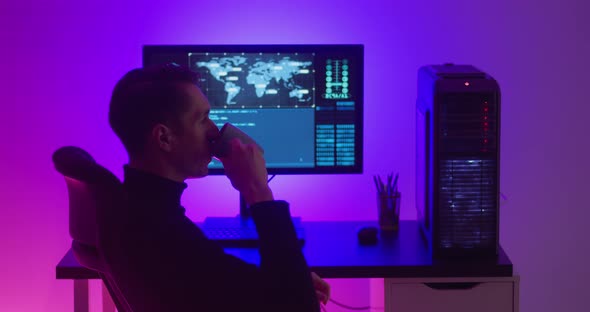 A Male Programmer Drinks Coffee While Working at a Computer in a Data Center at Night