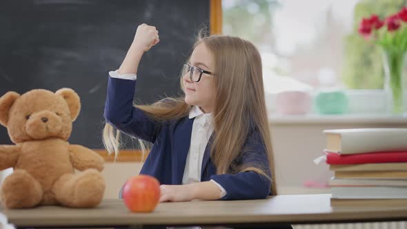 Positive Cute Schoolgirl Gesturing Strength Gesture Smiling Sitting at Desk with Teddy Bear and