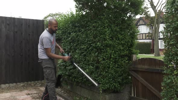 UK Asian Adult Bald Male Using Electric Hedge Trimmer Outside In Front Garden. Locked Off