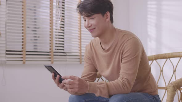 In his living room, an Asian man is on the phone, listening and conversing on his smartphone.