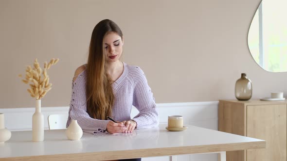 Cute Young Woman Sits Behind The Table And Signs Greeting Postcard