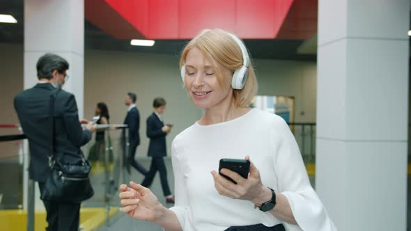 Attractive Mature Businesswoman in Headphones Walking and Dancing in Lobby Holding Smartphone