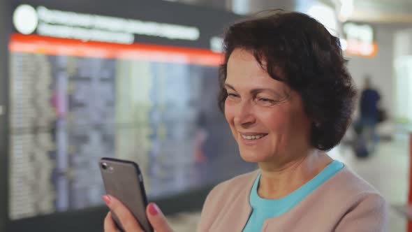 Smiling Aged Lady Standing in Airport and Checking Messenger on Phone