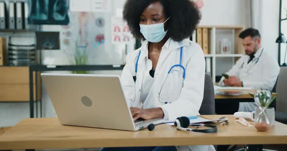 Black-Skinned Female Doctor in Facial Mask Sitting at the Table in front of Laptop and Typing