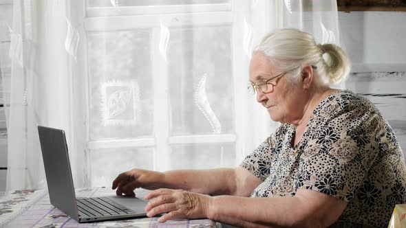 Gray-haired Woman Puts on Glasses and Types on the Laptop on the Table Against the Background