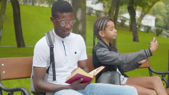 Absorbed African American Man Reading Book in Sunny Park As Little Girl Taking Selfie at the