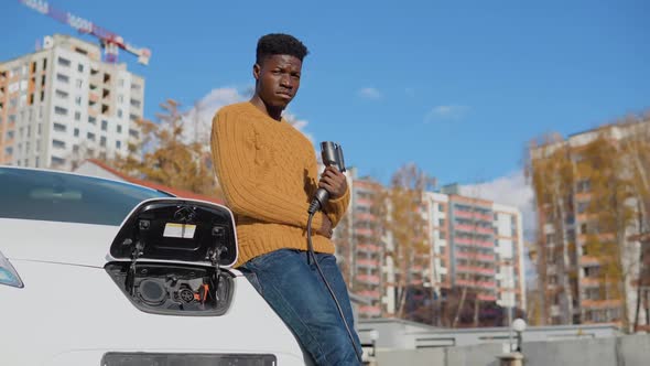 A Darkskinned Male Driver Holds a Charging Cable for an Electric Car Standing Near Opened Battery of