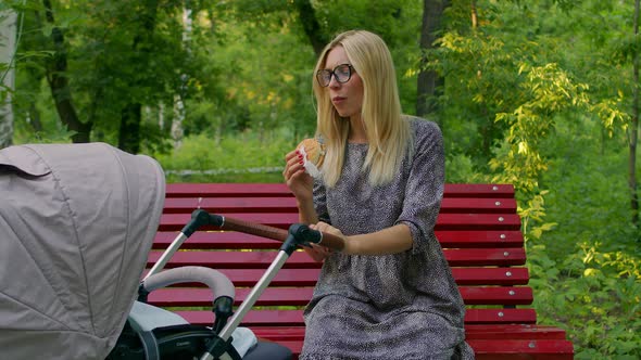 A Beautiful Mother Is Rocking a Stroller with a Newborn Baby and Eating a Burger. A Girl Is Sitting