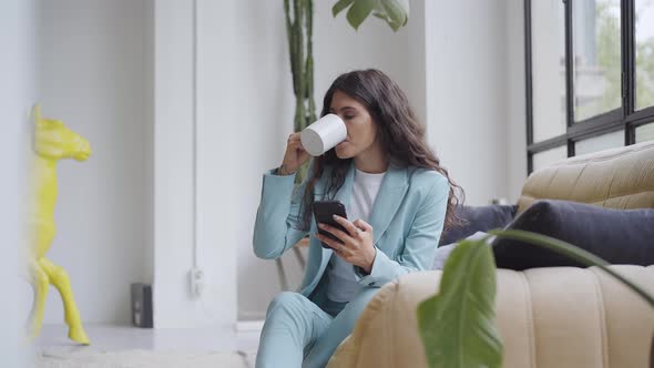 Beautiful Latina Businesswoman Drinking Coffee at Home Using a Smartphone Enjoying Relaxed Browsing