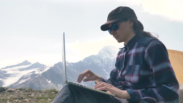 Lady Sits on Hill Top and Types on Laptop Against Mountains