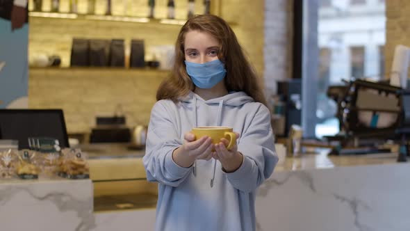Charming Young Woman in Coronavirus Face Mask Posing with Coffee Cup in Cafe
