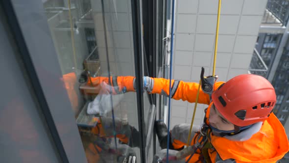 Equipped Window Washer in Helmet and Overalls Washes Glass with Screed at High Altitude of Building