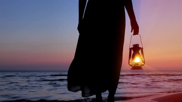 Silhouette of Woman in Dress Holding Kerosene Lantern on Seacoast at Sunset Sky Background