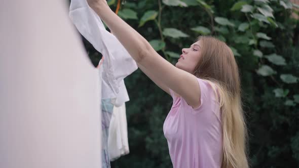 Side View Portrait of Young Woman Hanging White Clean Laundry on Rope Looking at Camera