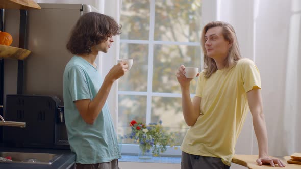 Smiling Young Gay Couple Drinking Coffee and Talking Together in t Kitchen in Morning