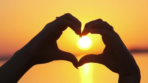 Silhouette of Female Hands Holding Sign in the Shape of a Heart at Orange Sunset