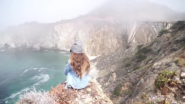 A young woman sitting on top of a cliff with the Bixby Creek Bridge in the background.