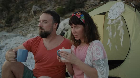 Young pretty woman and man drinking tea on pebble beach near the sea on sunset