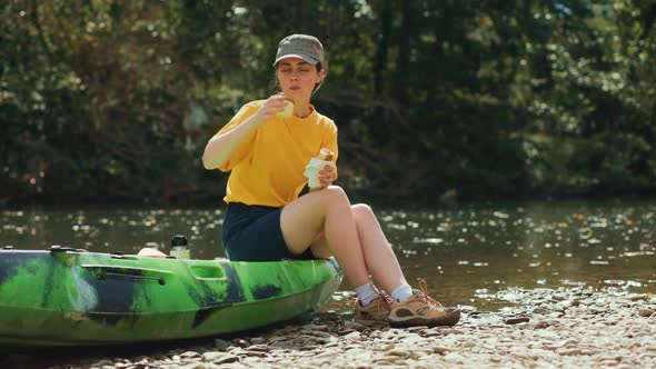 A young woman is sitting on a kayak and eating a hot dog, washing down a drink from a thermos.