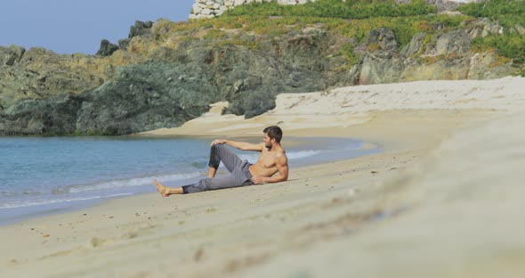 The Handsome Man with a Perfect Athletic Body in Gray Trousers Sits on a Deserted Beach in the