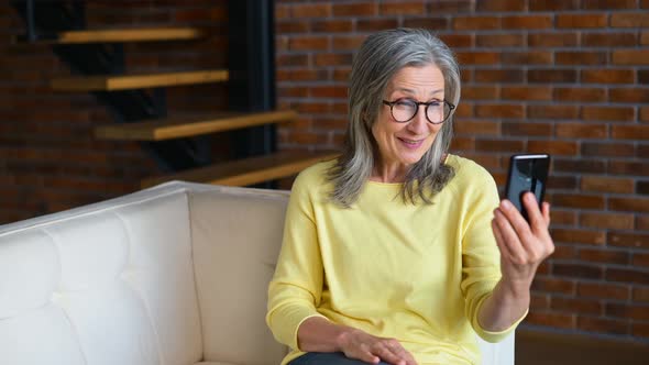 Positive Senior Mature Lady in a Yellow Jumper Talking Online on the Phonevideocalling