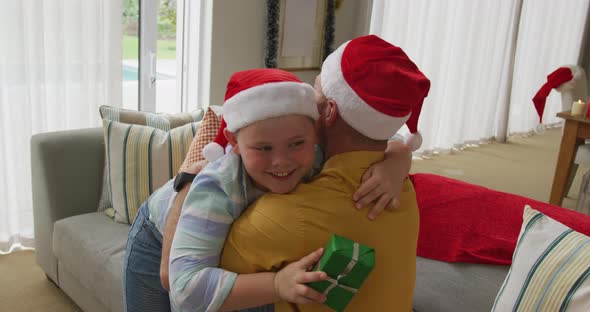 Caucasian boy wearing santa hat holding a christmas gift hugging his father at home