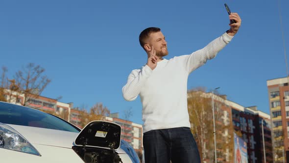 A Driver is Photographed on a Smartphone Camera Standing Near a White Electric Car