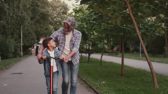 Grandfather and Grandson Riding a Scooter Walking in the Green Park