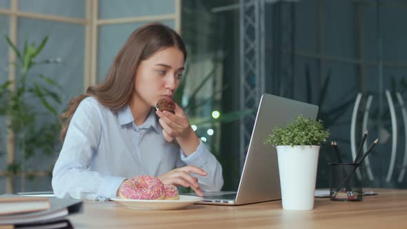 Young Businesswoman Eating a Glazed Donut While Working on Laptop in Office