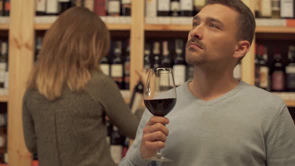 Handsome Pensive Guy with a Wine Glass in a Liquor Store. Woman in the Background Is Choosing a Wine