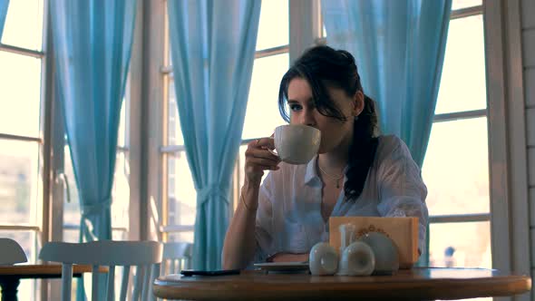 Girl Sits in A Restaurant at A Table with Large Panoramic Windows and Drinks Coffee 