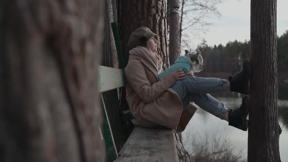 Young Woman in Coat is Sitting on Bench in Pine Forest with Dog on Her Lap Enjoying Nature