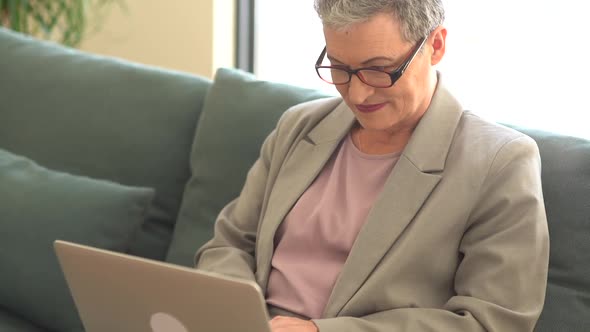 Active Happy Senior Woman Wearing Glasses and Short Haircut Sits on Sofa and Works Remotely Using