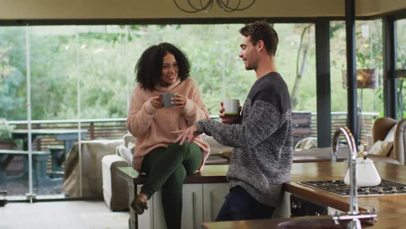 Happy diverse couple talking and drinking coffee in kitchen, woman sitting on counter