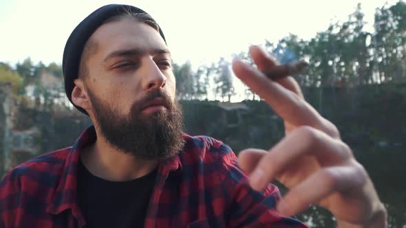 Portrait of a Bearded Guy Sitting in the Forest Near River and Smoking Brown Cigar.