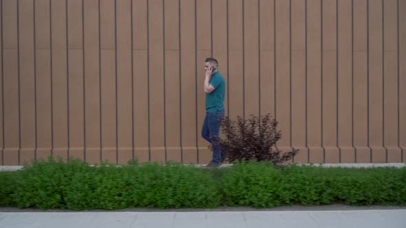 A Young Man Is Walking and Talking on the Phone. A Man Walks Near the Building and Talks on the