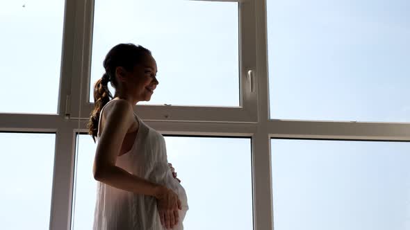 Pregnant Woman in White Dress Strokes Tummy Near Window