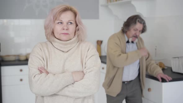 Scared Senior Caucasian Woman with Crossed Hands Listening To Angry Man Shouting and Hitting Table