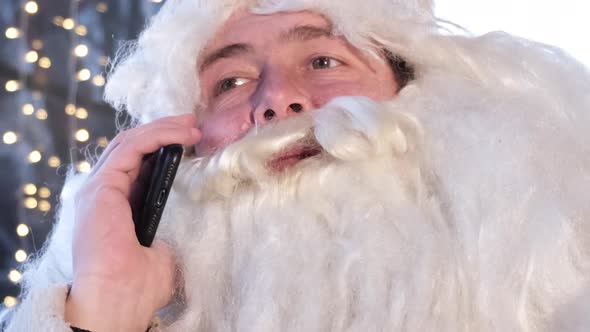 Santa Claus Uses a Smartphone While Sitting in a Cafe He is Resting and Talking on the Phone