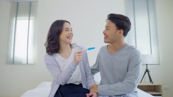 Asian happy couple, loving woman surprises boyfriend with good news to be future parents on bed.