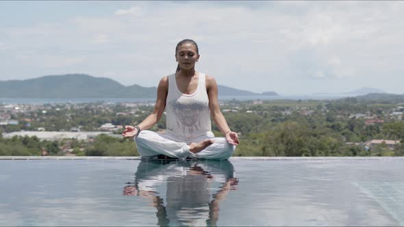 Calm Woman Practicing Yoga in Lotus Posture Poolside Against Resort Town