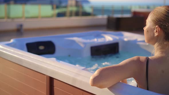 A Young Woman Relaxes in the Hot Tub on a Rooftop with a View on Mountains