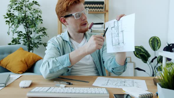 Joyful Man Architect Discussing Work on Video Call Talking Showing Blueprint Waving Hand Indoors at