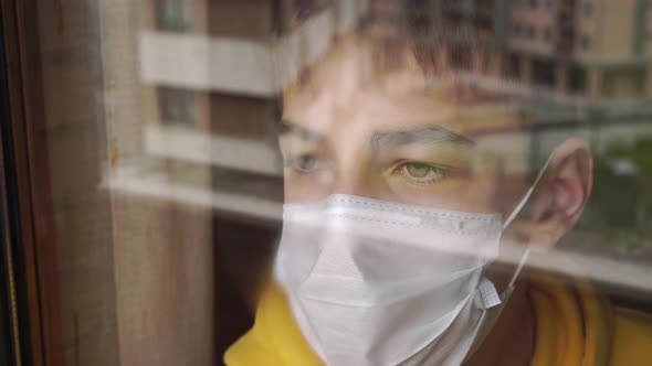Young man in a facial medical mask looks out of the window with sad eyes.