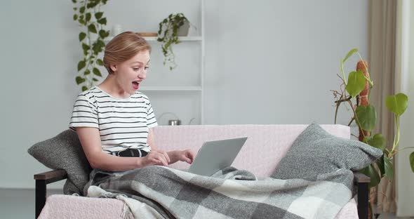 Carefree Caucasian Woman Sitting at Home on Sofa Covered with Blanket Looks at Laptop Plays Video